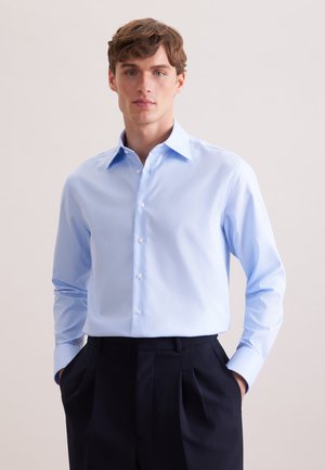 Młody mężczyzna o brązowych włosach, ubrany w jasnoniebieską koszulę na guziki, wpuszczoną w wysokie, czarne spodnie, z rękami w kieszeniach.