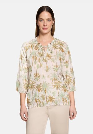 Vrouw draagt een lichtblouse met groene en bruine palmbomenprint en beige broek, staand tegen een effen achtergrond.
