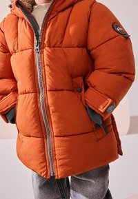 Doudoune orange avec capuche doublée de gris, fermeture éclair avant et poches à boutons-pression. Texture matelassée apportant chaleur et relief.