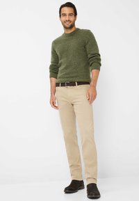 Maglione verde texturizzato, pantaloni beige, cintura marrone e scarpe scure. Il modello si trova su uno sfondo neutro, mostrando l'insieme dell'abbigliamento.
