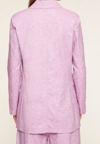 Světle růžové sako s texturovanou látkou, vybavené klopovým límcem, jedním zadním rozparkem a dvěma bočními kapsami. Střih na míru a dlouhé rukávy.