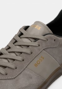 Gros plan d'une basket en daim gris avec des lacets gris et un logo doré "BOSS" sur le côté et la languette.