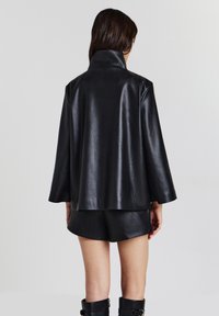 Persona che indossa una giacca nera in similpelle con colletto alto, pantaloni corti coordinati e stivali neri alti, vista di spalle su uno sfondo semplice.