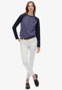 Kobieta o długich włosach, ubrana w niebieską koszulę z długim rękawem typu raglan, białe obcisłe jeansy z brązowym paskiem oraz ciemnobrązowe mokasyny, stojąca z nogami skrzyżowanymi.