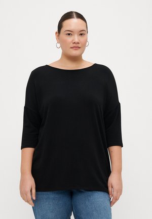 T-shirt nera oversize con maniche a tre quarti, scollo rotondo e una texture di stoffa liscia. Indossata con jeans blu chiaro.