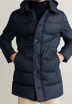 Homme portant un manteau d'hiver matelassé bleu foncé avec boutons et fermeture éclair, mains dans les poches latérales, pantalon beige visible en bas.