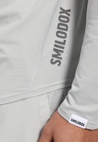 Hellgraues Sportshirt aus strukturiertem Stoff mit einem auffälligen "SMILODOX"-Logo in Dunkelgrau an der Seite und einem Etikett am Bündchen.