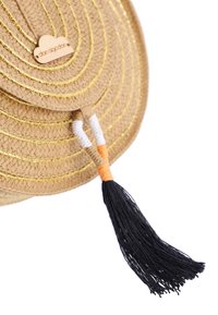 Bolso redondo de paja en color natural, con detalles dorados, un borlas decorativa y una etiqueta de madera con el logo. Textura trenzada prominente.