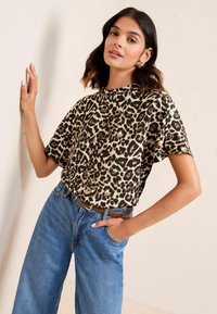 Top in stampa leopardata con maniche corte realizzato in tessuto leggero, abbinato a jeans blu in denim a vita alta e una cintura marrone, su uno sfondo neutro.