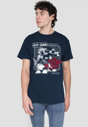 Junger Mann trägt ein blaues T-Shirt mit weißem Robotergrafik und rotem Text "Du hast 20 Sekunden, um zu gehorchen," kombiniert mit blauen Jeans.