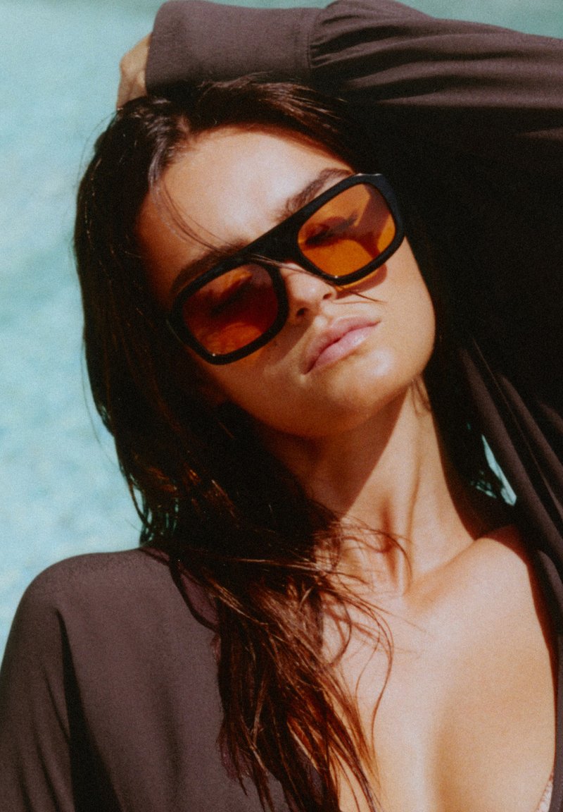 Femme aux longs cheveux foncés portant de grandes lunettes de soleil teintées orange et un haut noir, posant avec un bras levé derrière la tête.