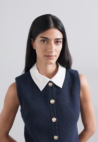 Robe sans manches bleu marine foncé avec un col blanc, des boutons dorés et un tissu texturé avec une silhouette ajustée.