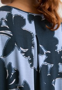 Close-up di una camicia con motivo floreale blu e nero, vista dal retro, con un dettaglio plissettato e capelli rossi visibili sul lato destro.