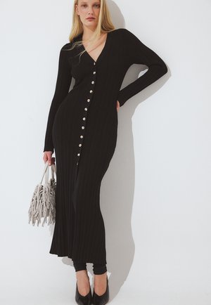 Kvinde iført en lang sort ribbet kjole med knapper, der holder en beige håndtaske med frynser, stående mod en hvid væg.