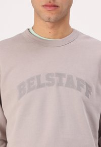 Felpa grigia chiara con scollo a girocollo, caratterizzata da una scritta ricamata "BELSTAFF" sul petto di una tonalità leggermente più scura. Texture morbida.