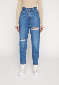 Jeans en denim bleu taille haute avec des déchirures sur les genoux, délavage clair, design classique à cinq poches et coupe fuselée.