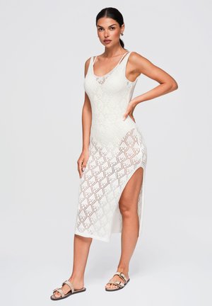 Femme aux cheveux foncés vêtue d'une longue robe blanche sans manches en maille avec une fente latérale, portant des sandales beiges, debout devant un fond clair uni.