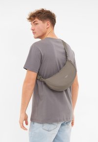 Olive green sling bag worn diagonally across the back, made from smooth material with a curved shape, featuring a single adjustable strap.