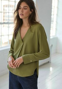 Blouse olive vert, à manches longues, avec un décolleté en V et des fentes sur les côtés. Présente une texture lisse et de petits boutons argentés décoratifs sur les manches.