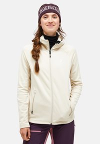 Cream-colored softshell jacket with a high collar, zipper pockets, and a fitted design. Paired with a dark headband, features a logo.