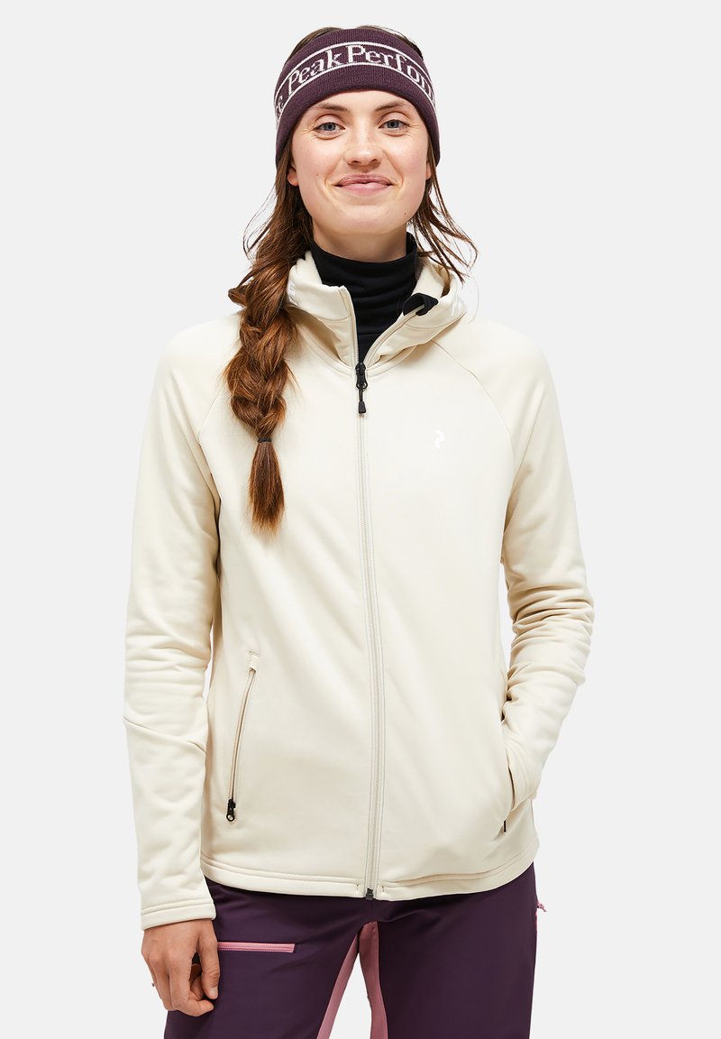 Cream-colored softshell jacket with a high collar, zipper pockets, and a fitted design. Paired with a dark headband, features a logo.