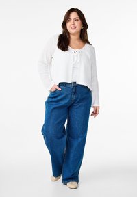Cardigan bianco in maglia texturizzata con chiusura a fiocco, indossato sopra una maglietta bianca, abbinato a jeans blu larghi e scarpe beige slip-on.