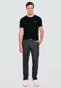 Černé bavlněné tričko s červeným textem, kombinované se šedými kostkovanými kalhotami. Model má na sobě bílé tenisky a hnědý pásek, stojí před šedým pozadím.