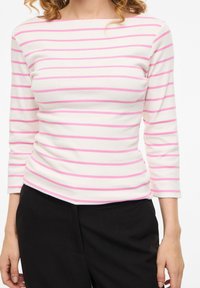 Γυναίκα που φοράει λευκό μακρυμάνικο τοπ με ροζ οριζόντιες ρίγες και μαύρο παντελόνι, στέκεται μπροστά σε μια απλή φόντο.