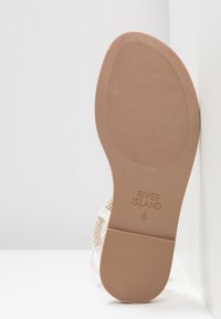 Semelle d'une sandale plate beige avec grip texturé, marque River Island, taille 4, inclinée contre un mur blanc et une surface.