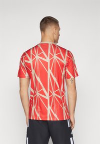Camiseta atlética roja con un patrón geométrico beige, de manga corta y cuello redondo, fabricada con un material suave y ligero.