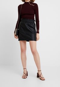 Minifalda de imitación cuero negra con cinturilla elástica, combinada con un suéter de cuello alto acanalado burdeos con botones metálicos y mangas largas.