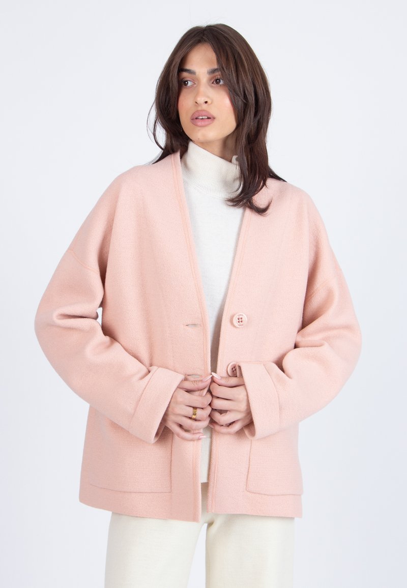 Vrouw in een lichtroze, geknoopte wollen jas over een witte coltrui en crèmekleurige broek, die iets opzij kijkt.