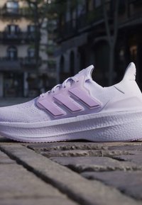 Бял и светлолилав маратонка Adidas на каменна настилка с размазани градски сгради на заден план.