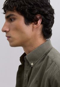 Olive green corduroy shirt with a button-down collar and a subtle logo on the chest. Features fine ribbed texture and contrasting buttons.