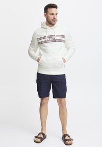 Sudadera blanca con capucha y rayas horizontales rojas y azul marino, combinada con pantalones cortos cargo azul marino y sandalias marrones. Presenta un bolsillo frontal.