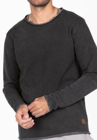 Langzaamslavende shirt in donkergrijs, gemaakt van zachte stof. Heeft een brede halslijn, ruwe randen en een klein merkembleem aan de onderkant.