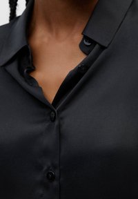 Chemise en satin noir avec un col pointu, présentant une texture lisse et des boutons noirs. Le tissu a une légère brillance et un design épuré et minimaliste.
