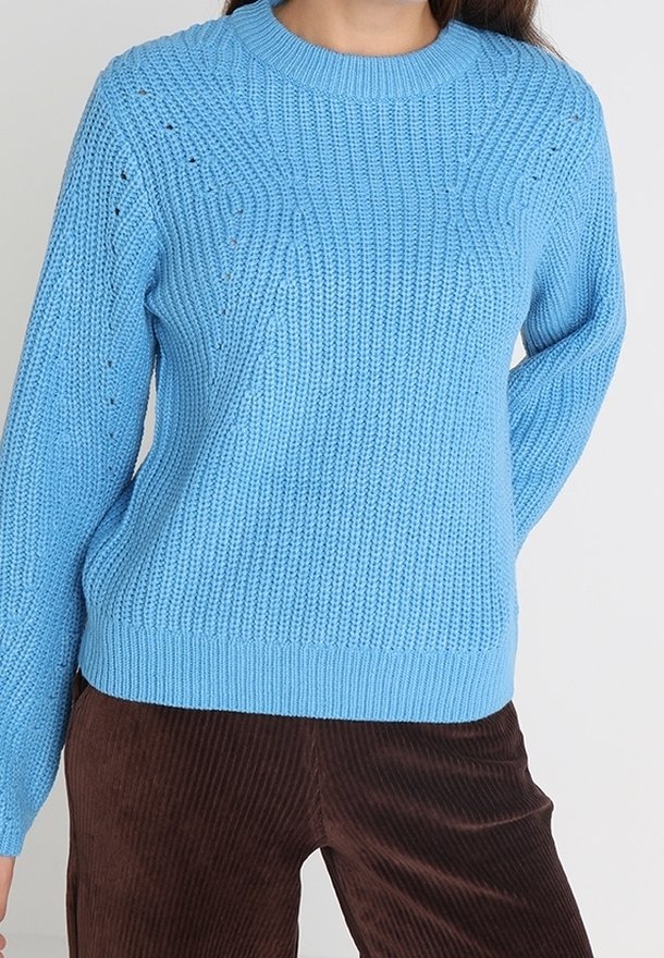 Femme portant un pull en tricot bleu clair associé à un pantalon en velours côtelé marron foncé, debout avec une main derrière le dos.