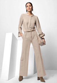 Beige blouse met een capuchon en knoopsluiting, gecombineerd met beige wijde broekspijpen met cargozakken, en aangevuld met een kleine handtas.