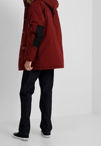 Rode gevoerde parka-jack met een bont gevoerde capuchon, zwarte accenten op de mouwen en drukknoopsluitingen, gecombineerd met donkere jeans en zwarte sneakers.