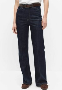 Donkerblauwe spijkerbroek met rechte pijpen, hoge taille en een bruine leren riem. Zichtbare stiksels en klassieke vijfzakkenontwerp.