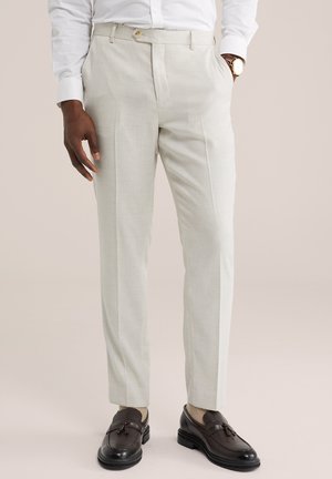 Homme portant un pantalon beige clair sur mesure, une chemise blanche, une montre marron clair, des chaussettes beiges et des mocassins à pampilles marron foncé. Mains détendues le long du corps.