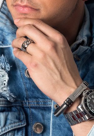 Silver metal ring with a chain design, black textured bracelet, and a silver watch with a black face, worn on a hand against a denim background.