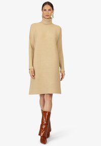 Robe beige à col roulé côtelé, à manches longues, coupée au niveau des genoux et à la coupe décontractée ; assortie à des bottes montantes en cuir marron brillantes avec embossage croco.