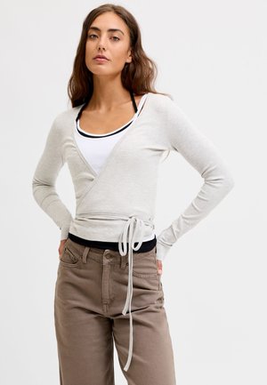 Femme aux longs cheveux bruns portant un pull cache-cœur gris clair sur un débardeur blanc et un pantalon marron, debout, les mains sur les hanches, sur un fond uni.