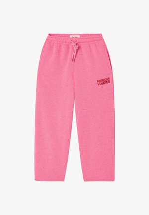 Osoba stojaca na svetlej podlahe pri jednofarebnej stene, oblečená v voľných ružových teplákoch so sťahovacou šnúrkou a béžových sandáloch s prackou.