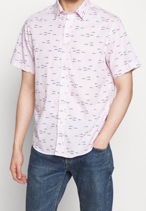 Mann trägt hellrosa kurzärmeliges Hemd mit kleinem schwarzem Fischmuster und blaue Jeans, eine Hand in der Tasche.