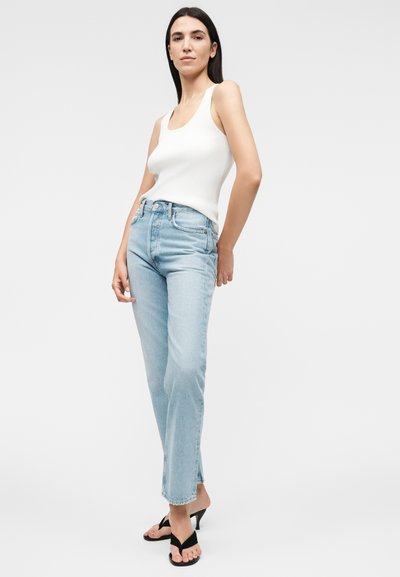 Γυναίκα με μακριά σκουρόχρωμα μαλλιά που φοράει λευκό αμάνικο τοπ, ανοιχτό μπλε τζιν με ψηλή μέση και μαύρα σανδάλια με τακούνι, στέκεται σε λευκό φόντο.