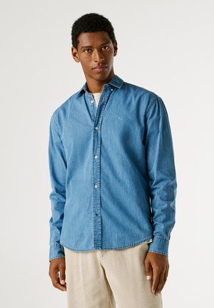 Jonge man die een lichtblauw denim blouse met knopen en beige corduroy broek draagt, staande tegen een effen witte achtergrond.