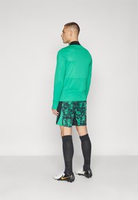 Grünes langärmliges Shirt mit schwarzem Kragen, kombiniert mit gemusterten grünen Shorts. Schwarze Kniestrümpfe und weiße Fußballschuhe mit gelben Akzenten.